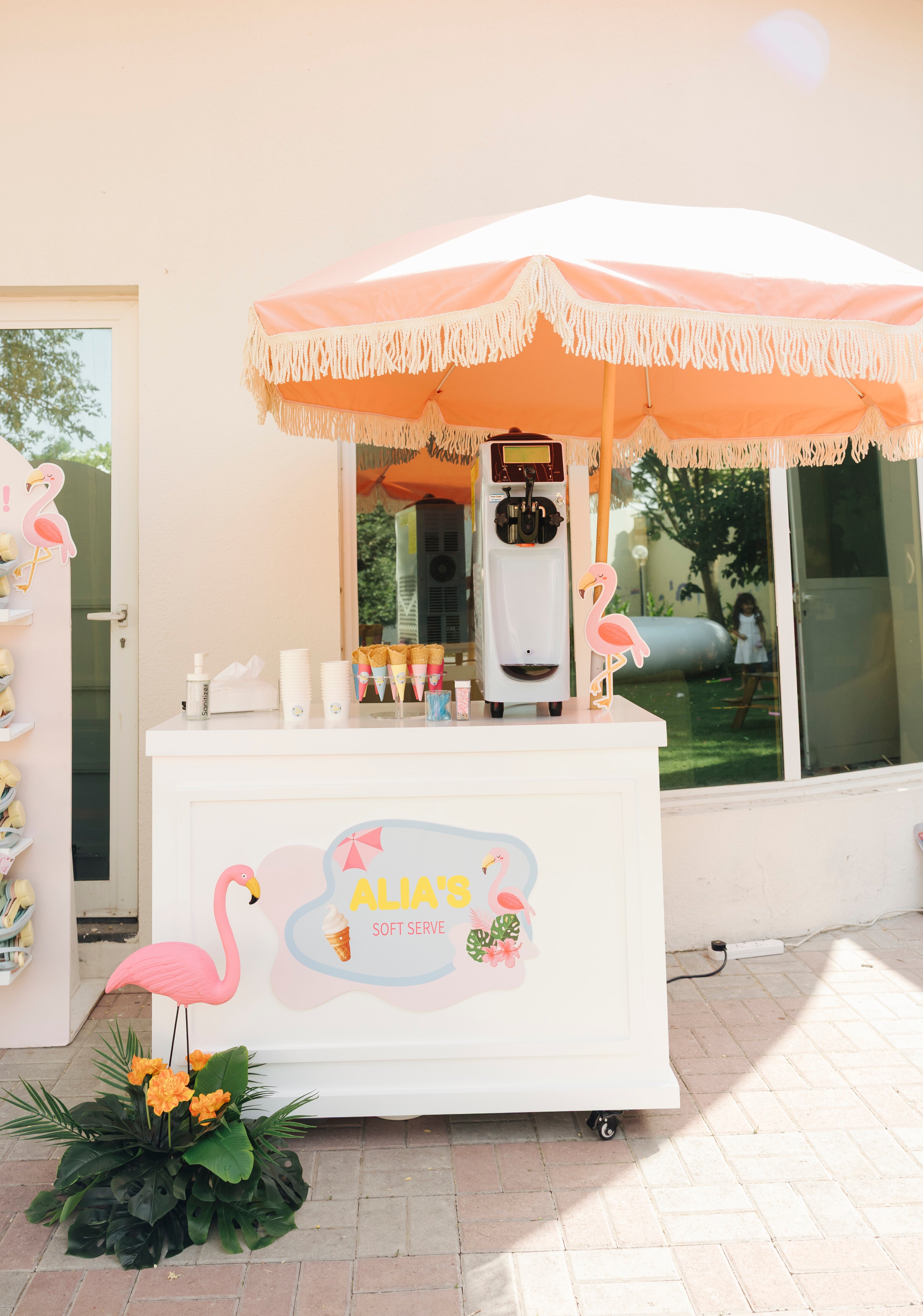 A white ice cream cart labeled "Alia's Soft Serve" with colorful cones, a soft serve machine, and pink flamingo decorations under a peach umbrella.