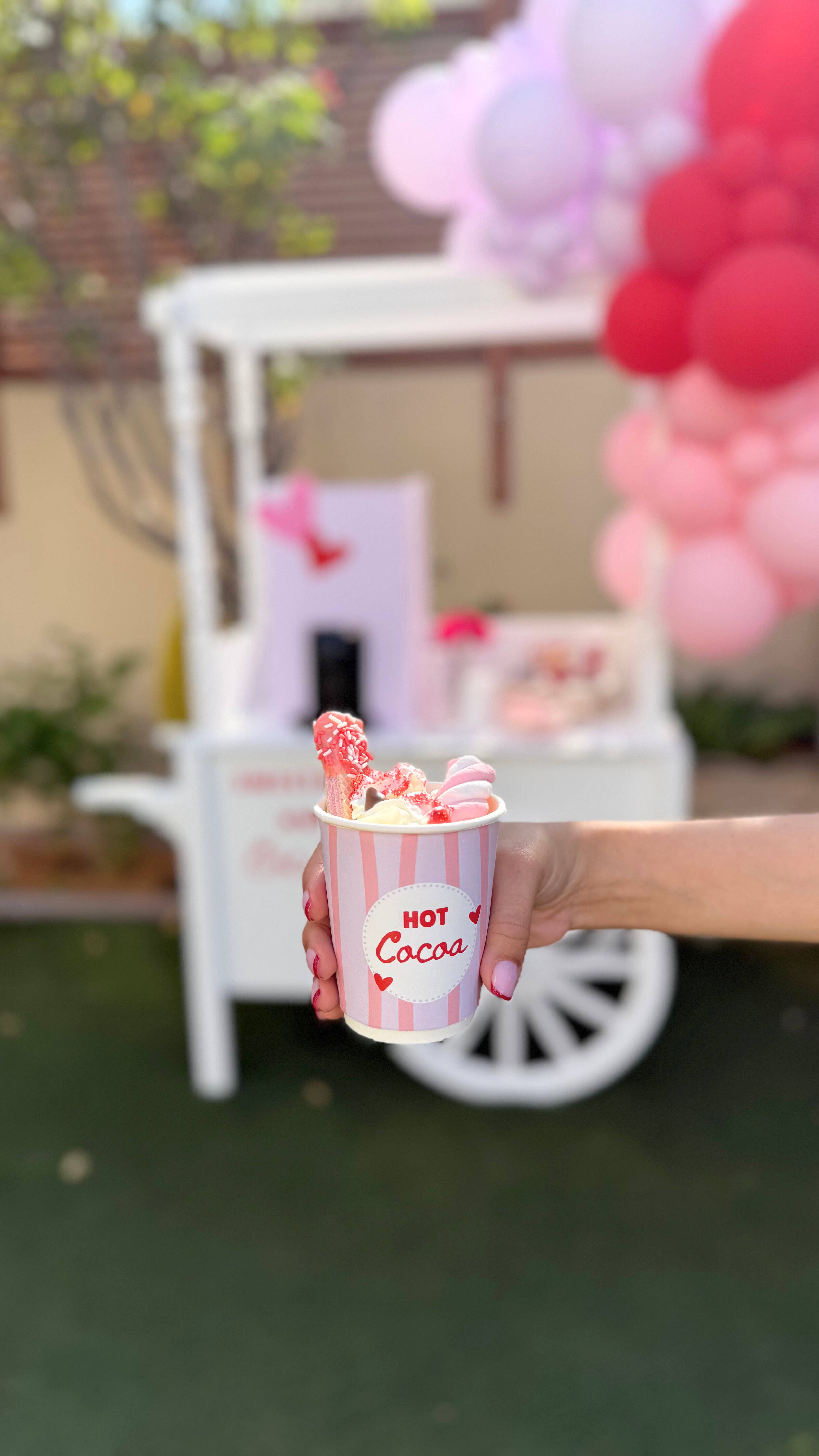 A hand holding a decorated cup of hot cocoa with pink toppings in front of a themed hot cocoa cart and pink balloon decor.