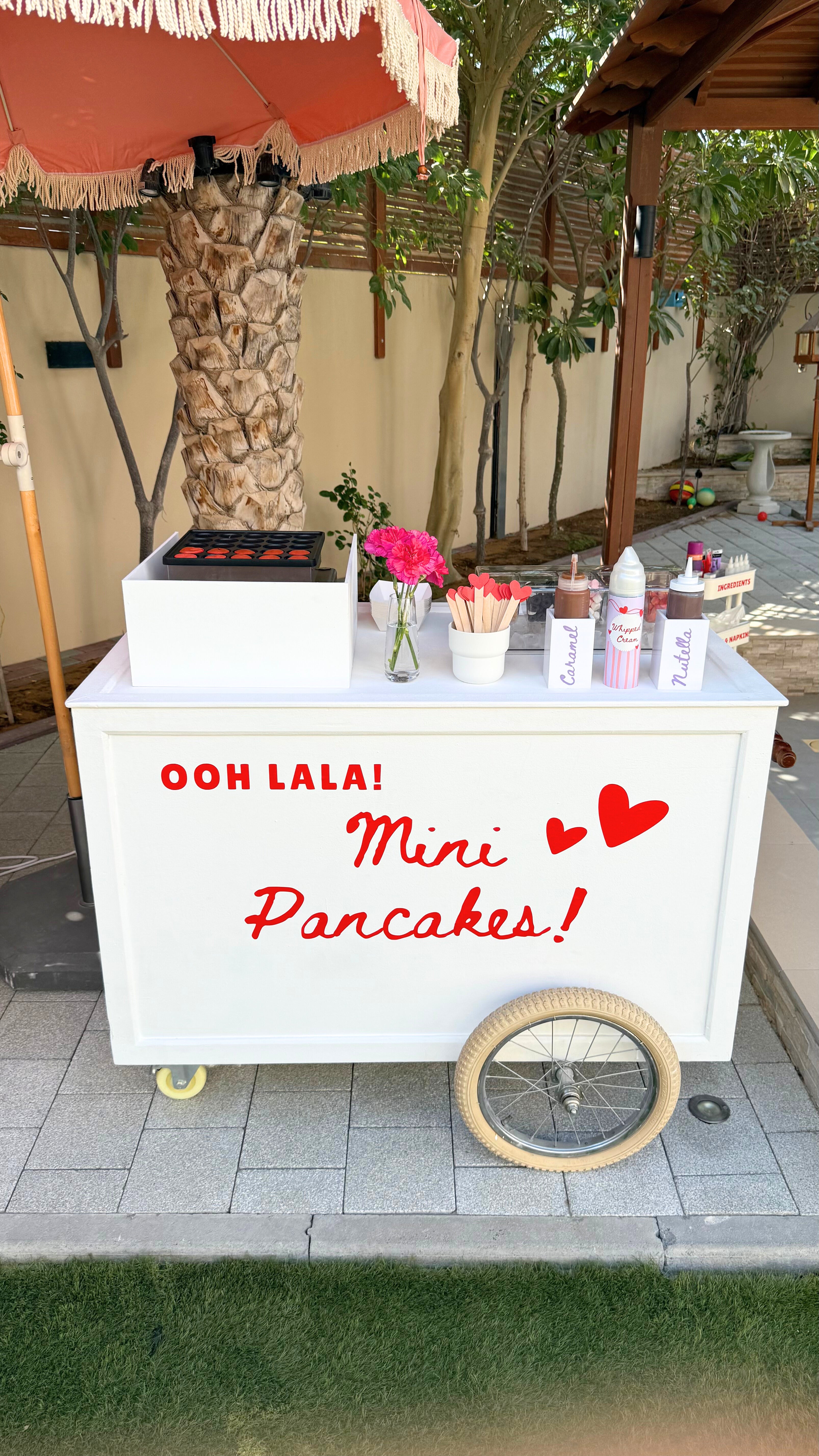 A decorated mini pancake cart with red text reading "Ooh Lala! Mini Pancakes!" featuring flowers, toppings, and syrup bottles under a fringed umbrella in a garden setting.