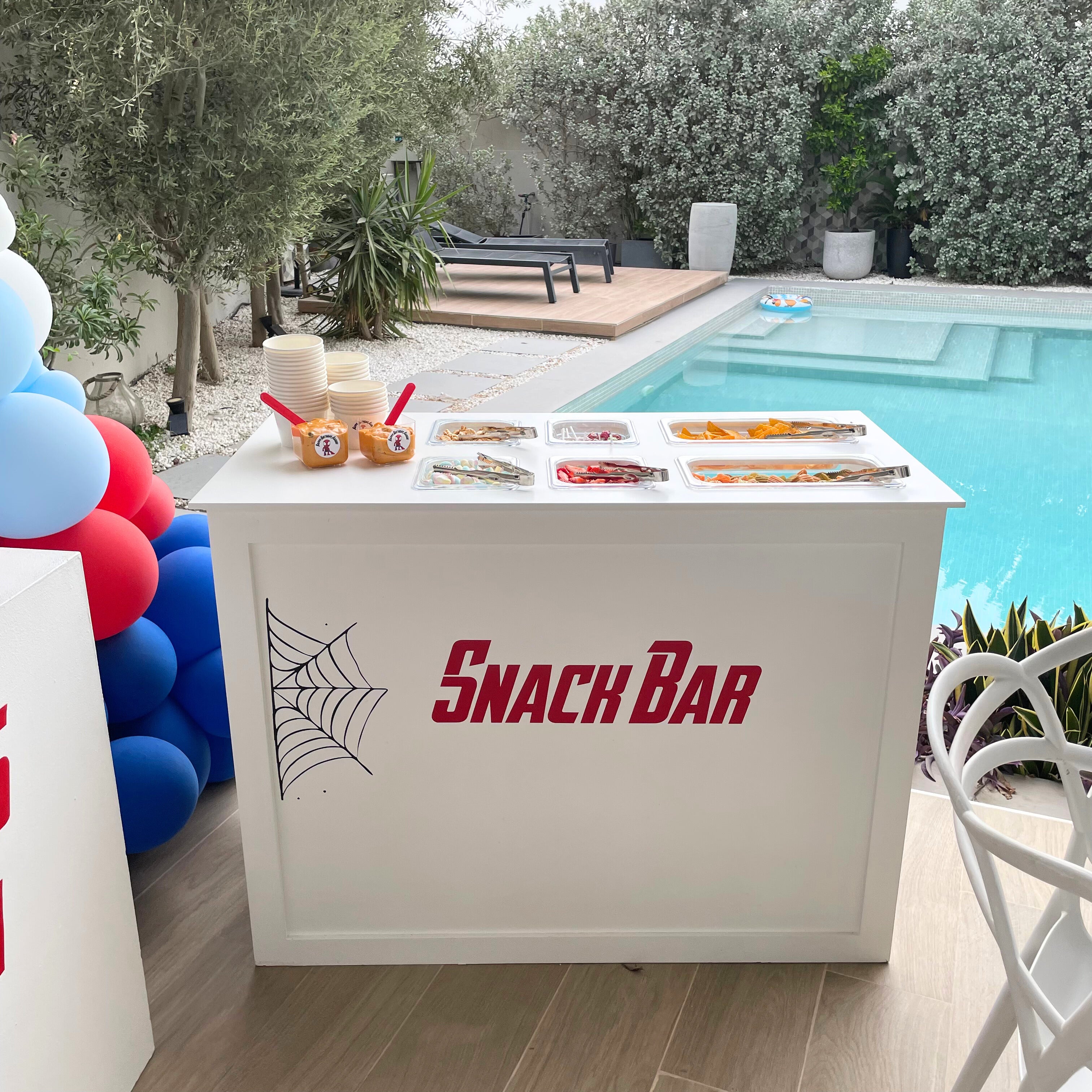 A white snack bar setup by a pool, featuring various treats in trays and surrounded by red and blue balloons.