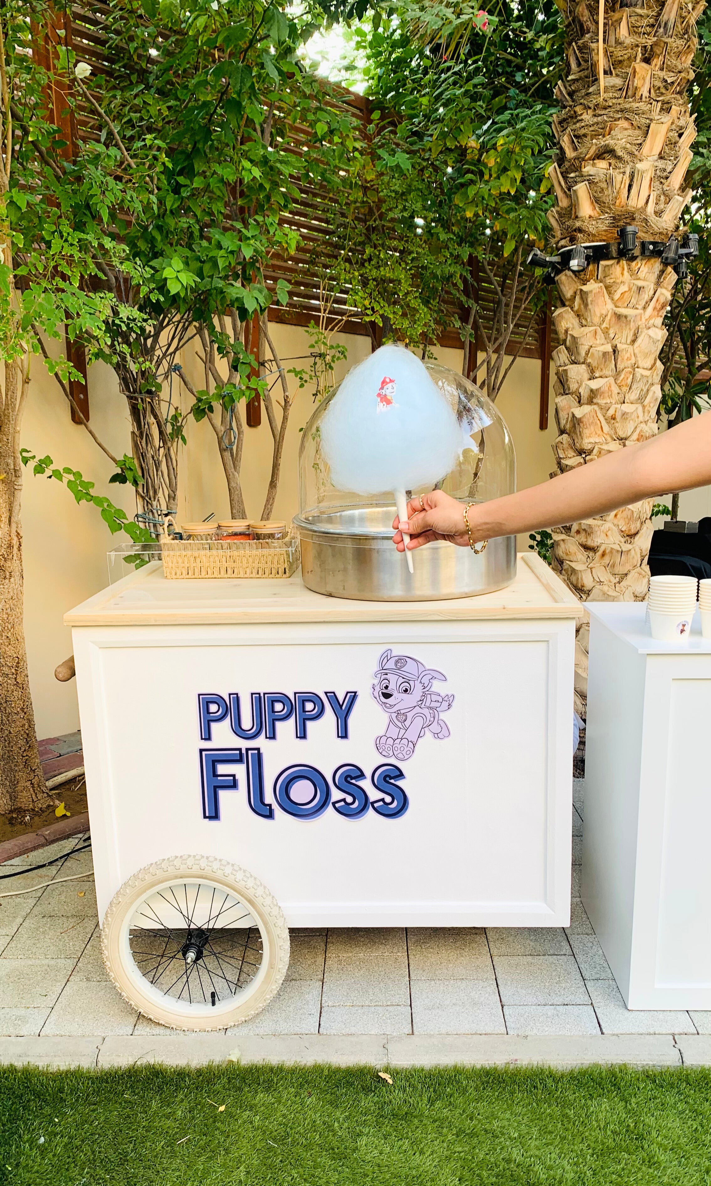 A white cotton candy cart labeled "Puppy Floss" with cartoon puppy art, set outdoors under trees. A hand is holding a blue cotton candy cone with a character printed on it.