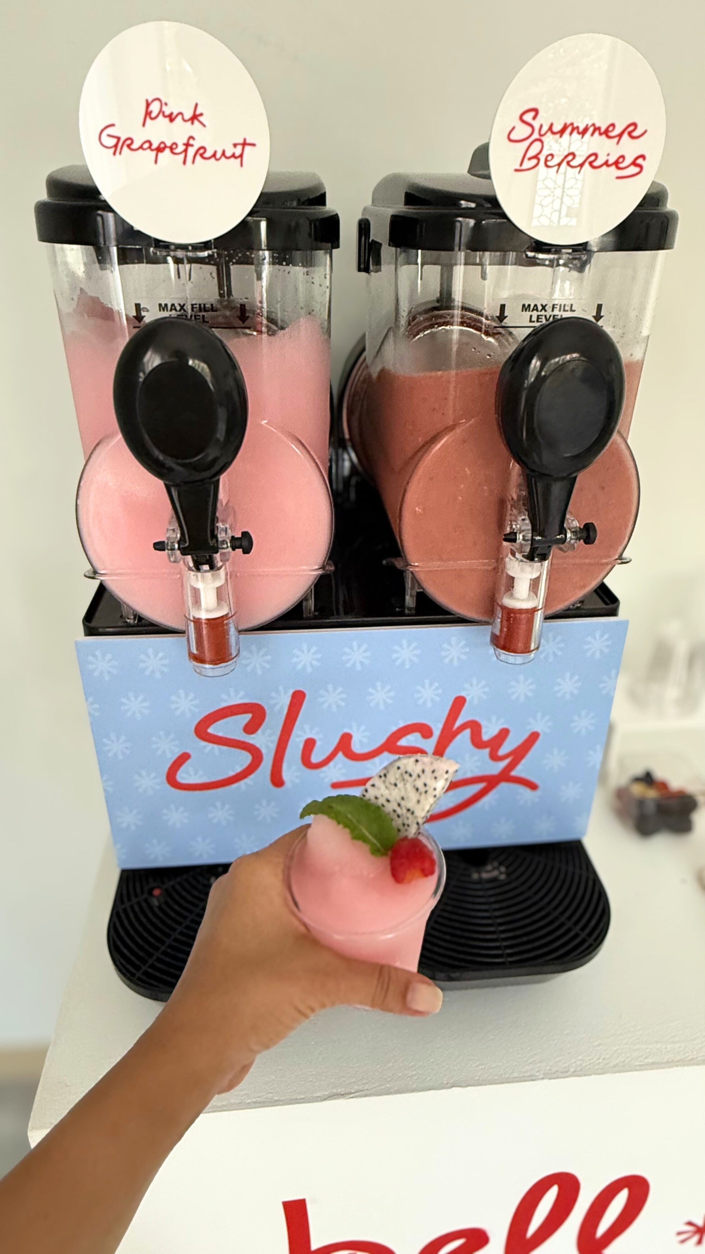 A twin slushy machine offering Pink Grapefruit and Summer Berries flavors, with a hand holding a pink slushy cup topped with fruit and mint. The label reads "Slushy" in red cursive.